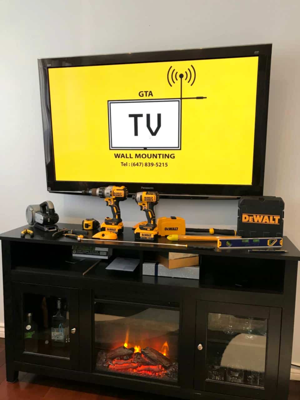 The GTA TV Wall Mounting tool kit laid out below a mounted Panasonic screen: hammer drill, impact driver, laser, levels and masonry bits