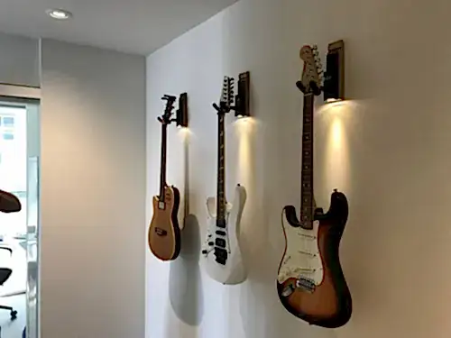 Three guitars hung on wall mounts under picture lights along an office corridor