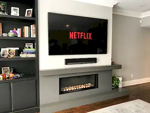 TV and soundbar mounted above a grey linear gas fireplace, with dark built-in shelving alongside