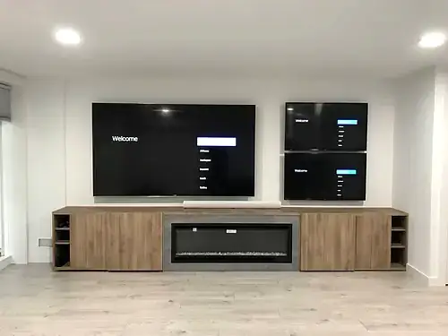 Three screens mounted together, one large and two stacked, above oak cabinetry with a linear fireplace