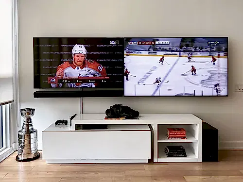 Two flat screens mounted side by side on a white wall above a low console, set up for two games at once