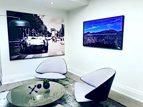 TV mounted in an office lounge corner beside a large canvas print, with two designer chairs