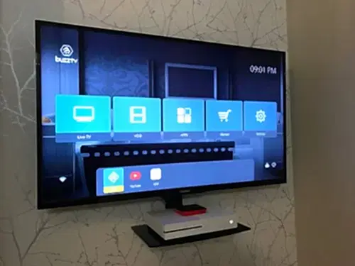 TV mounted on a patterned wallpaper wall above a small glass AV shelf holding a streaming box