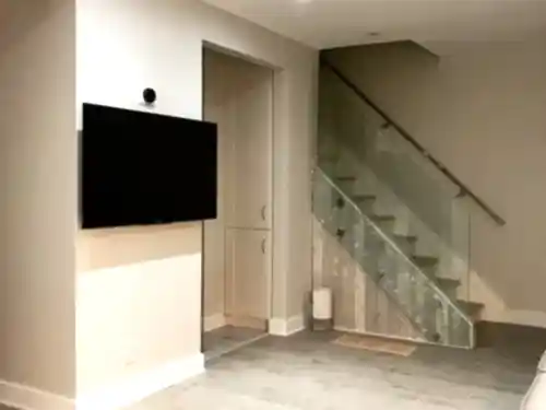 TV mounted on a basement half-wall beside a glass-railed staircase, with a sensor fitted above the screen