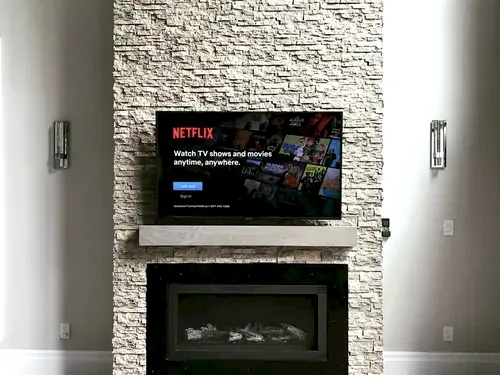 TV mounted on a stacked stone chimney above a timber mantel and linear fireplace, between two sconces