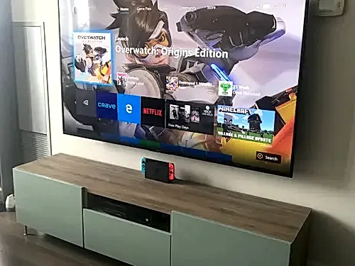 Large TV mounted on a white wall above a sage green and oak console with a games console below