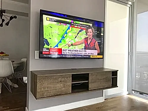 TV mounted on an angled condo wall above a grey floating console, with the dining area beyond