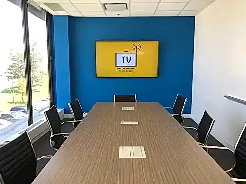 TV mounted on a blue boardroom wall, seen down the length of the table toward the window