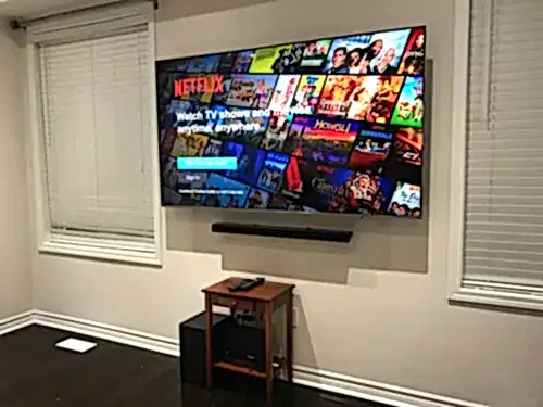TV and soundbar mounted on a grey wall between two windows, above a small wood side table