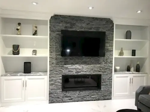 TV mounted on a grey stacked stone chimney above a linear fireplace, between marble-topped built-ins