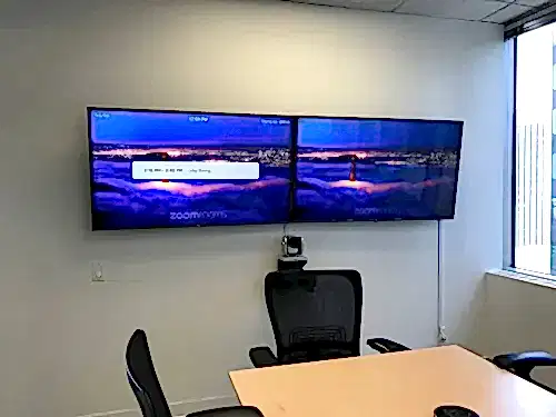 Two TVs mounted side by side in a meeting room with a conference camera fitted between them