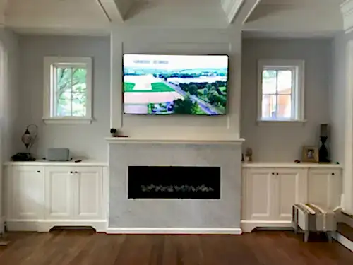 TV mounted above a marble fireplace surround and painted mantel, between white cabinets and two windows
