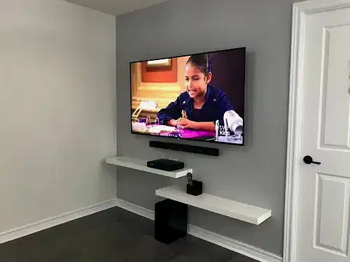 TV and soundbar mounted on a grey wall above two staggered white floating shelves and a subwoofer