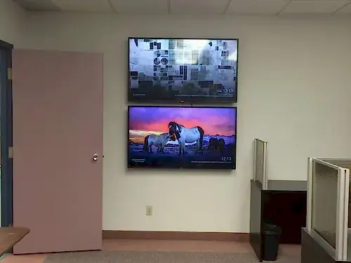 Two TVs stacked one above the other on an office wall beside workstation partitions