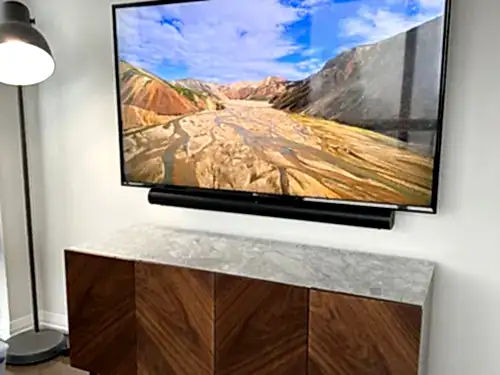 TV with a soundbar mounted below it on a white wall, above a marble-topped walnut credenza