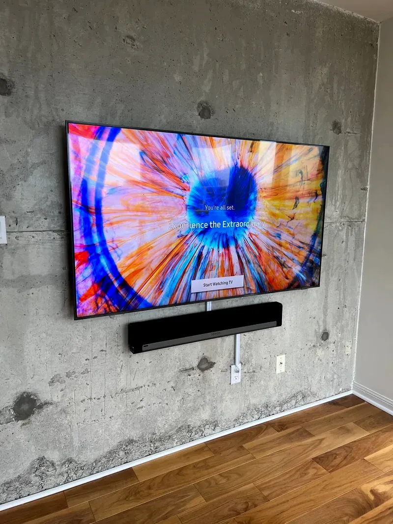 A flat-panel TV and soundbar surface-mounted on an exposed poured-concrete wall in a Toronto condo, with the power cable run to a visible outlet