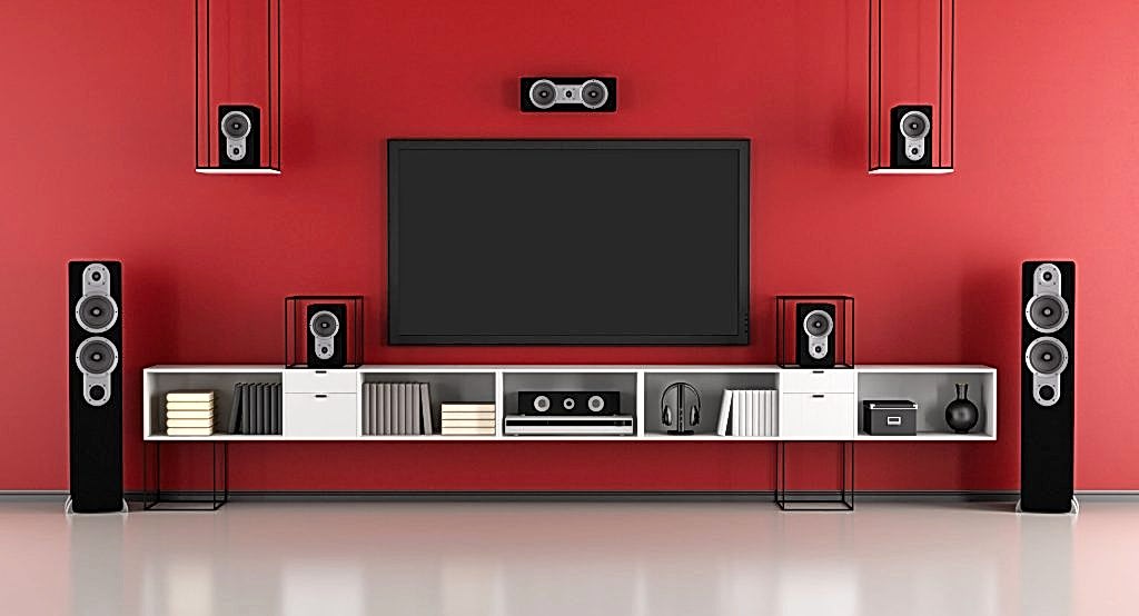 A modern home entertainment setup with a large flat-screen TV wall mounting on a red wall. Surrounding the TV, there are five black speakers: two tall floor-standing speakers, two smaller ones on shelves, and one center speaker above the TV. The shelves house books and decorative items.