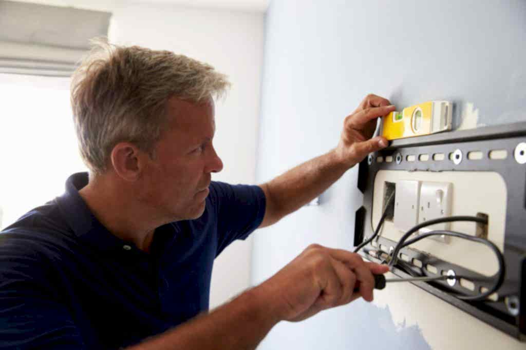 A man in a navy shirt uses a spirit level and screwdriver to install or adjust a TV wall mounting bracket. The surrounding area has a light blue wall with some paint patches. He is carefully aligning the unit to ensure it is level.