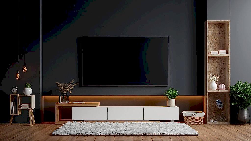 A modern, minimalist living room features TV Wall Mounting with a large flat-screen TV on a dark wall. Below, a white stand holds plants and decor. On the left, a small wooden side table boasts a lamp and clock, while to the right stands a tall wooden shelf adorned with plants and vases. A fluffy white rug covers the wooden floor.