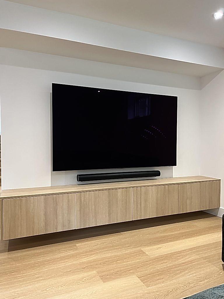Black soundbar mounted on the wall in the gap between a wall mounted TV and a floating oak cabinet